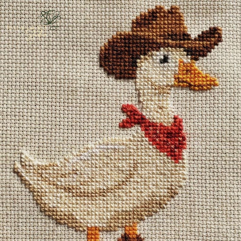 K&ouml;nnte beinhalten: Kreuzstich-Design einer Ente mit braunem Cowboyhut und rotem Halstuch. Die Ente ist in Beige-, Braun- und Oranget&ouml;nen auf einem neutralen Stoffhintergrund gestickt. Das Kunstwerk ist eine charmante, rustikale Darstellung.