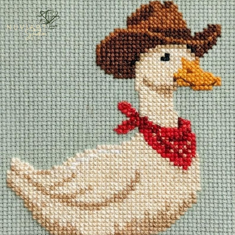K&ouml;nnte beinhalten: Kreuzstichbild einer Gans mit braunem Cowboyhut und rotem Halstuch. Die Gans hat einen gelben Schnabel und ist vor einem hellblauen Hintergrund platziert. Der Kreuzstich ist detailliert und strukturiert.
