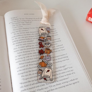 May include: A clear bookmark with fall-themed illustrations, including a sweater, pumpkin, and coffee cup. The bookmark is placed on an open book with text. A decorative pumpkin sign is in the background.