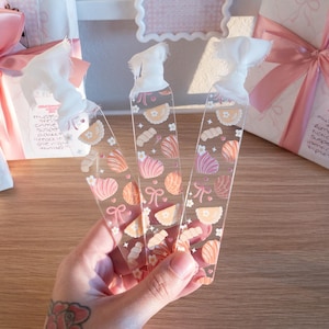 May include: Clear acrylic bookmarks with a seashell and bow pattern in pink, peach, and white are held in a hand. The bookmarks have a white fabric top. Gift boxes with pink bows and patterned paper are in the background.