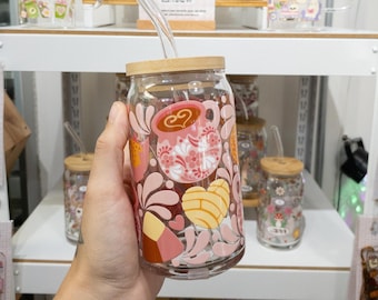 Op de afbeelding: Een doorzichtige glazen beker met een bamboe deksel en een glazen rietje, voorzien van een kleurrijke grafische print van roze koffiemokken, geel hartvormig concha-brood en abstracte bloemvormen in roze, oranje en witte tinten.
