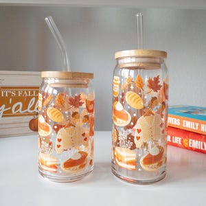 May include: Two clear glass tumblers with bamboo lids and clear straws. The tumblers are decorated with autumn-themed illustrations, including leaves, pumpkins, and a sweater. The tumblers are on a white shelf.