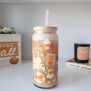 May include: A glass tumbler with a bamboo lid and a clear straw, filled with a creamy beverage. The tumbler is decorated with fall-themed stickers, including pumpkins, leaves, and a sweater. A wooden box with the text "Fall" and a pumpkin is in the background.
