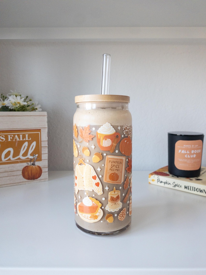 May include: A glass tumbler with a bamboo lid and a clear straw, filled with a creamy beverage. The tumbler is decorated with fall-themed stickers, including pumpkins, leaves, and a sweater. A wooden box with the text "Fall" and a pumpkin is in the background.