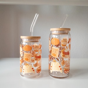 May include: Two clear glass tumblers with bamboo lids and glass straws. The tumblers are decorated with fall-themed illustrations, including pumpkins, recipe cards, and sweaters. The tumblers are of different sizes and are displayed on a white surface.
