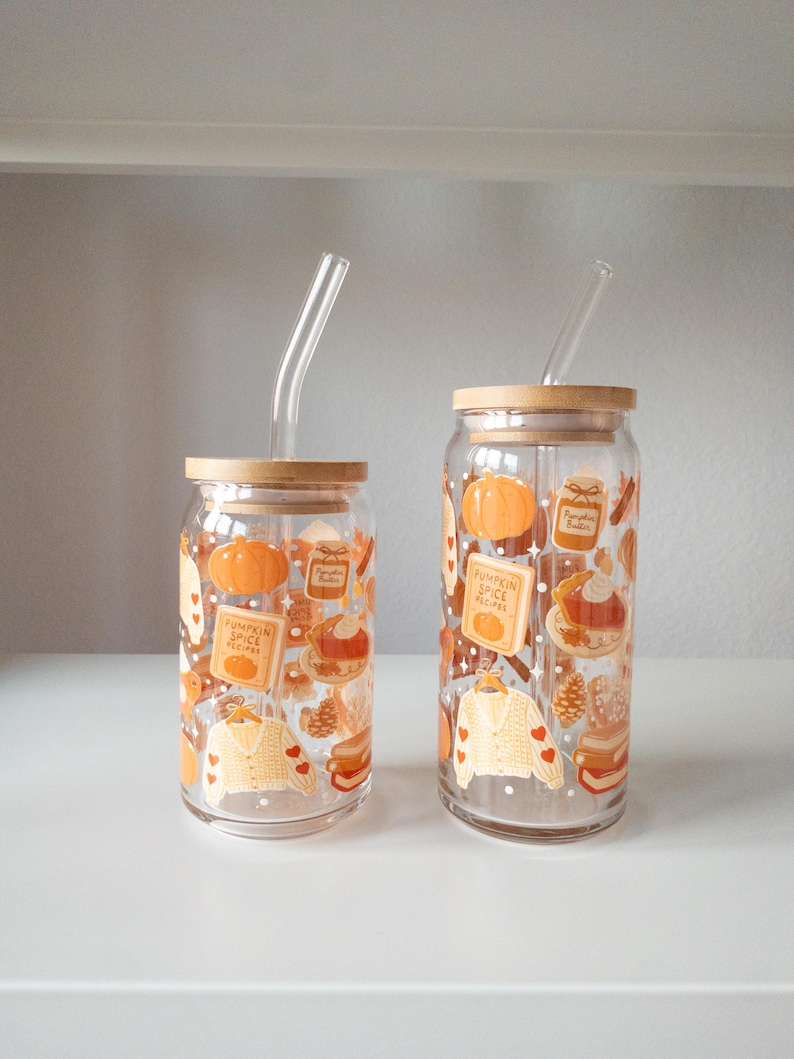 May include: Two clear glass tumblers with bamboo lids and glass straws. The tumblers are decorated with fall-themed illustrations, including pumpkins, recipe cards, and sweaters. The tumblers are of different sizes and are displayed on a white surface.