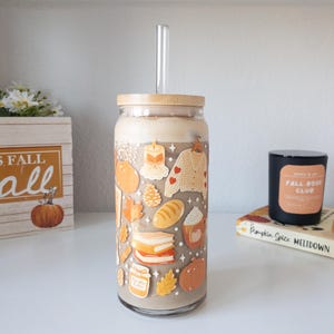 May include: A glass tumbler with a bamboo lid and a clear straw, filled with a creamy beverage. The glass is decorated with fall-themed stickers, including pumpkins, leaves, and a sweater. A wooden box with the text "FALL" is in the background.