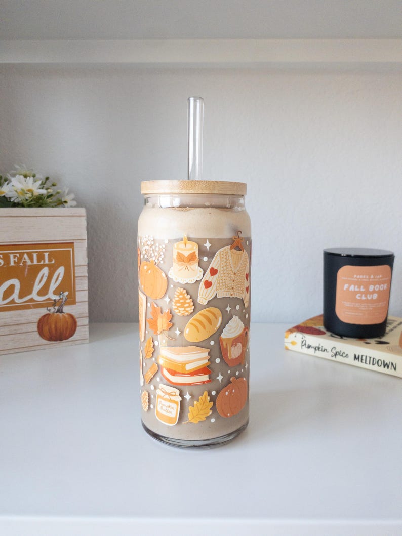 May include: A glass tumbler with a bamboo lid and a clear straw, filled with a creamy beverage. The glass is decorated with fall-themed stickers, including pumpkins, leaves, and a sweater. A wooden box with the text "FALL" is in the background.