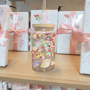 May include: A clear glass tumbler with a wooden lid and a glass straw. The tumbler is decorated with a colorful floral and fantasy design, including books, mushrooms, and an owl. The tumbler is displayed on a wooden shelf.