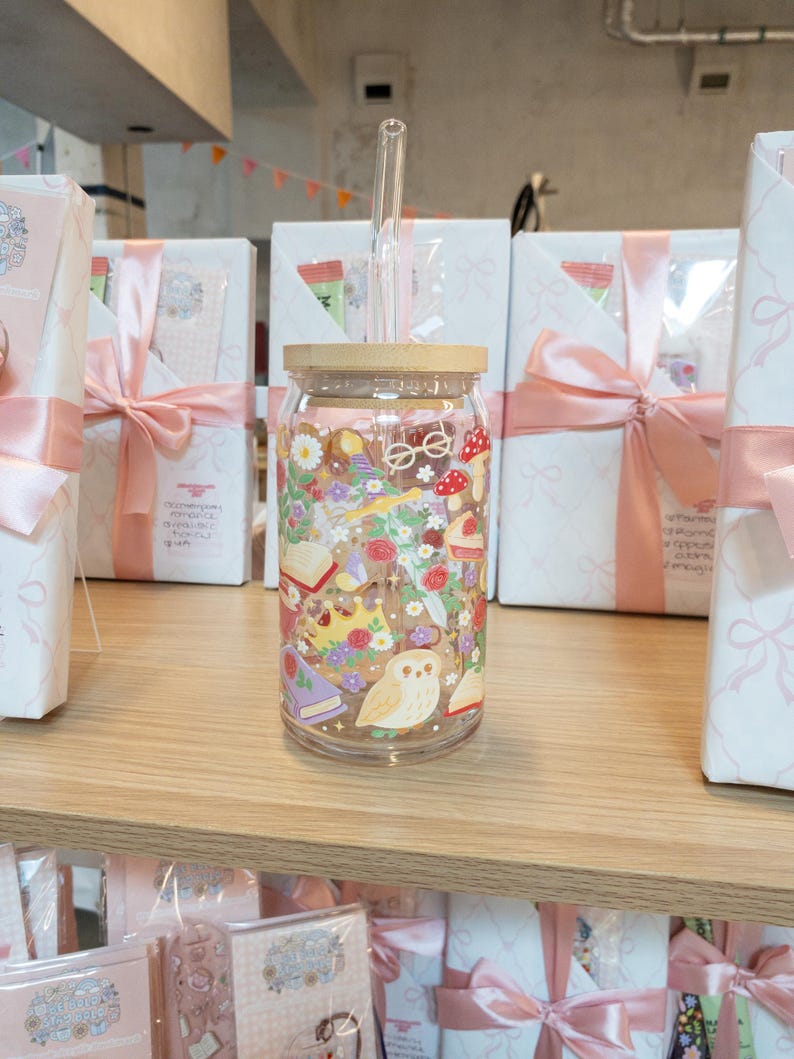 May include: A clear glass tumbler with a wooden lid and a glass straw. The tumbler is decorated with a colorful floral and fantasy design, including books, mushrooms, and an owl. The tumbler is displayed on a wooden shelf.