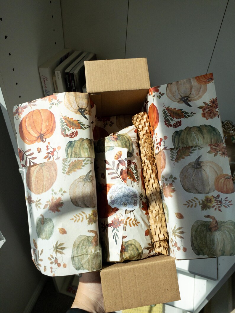 May include: An open cardboard box with autumn-themed wrapping paper featuring pumpkins and fall foliage. Inside, wrapped gifts are visible, protected by brown paper packaging. The box is held by a person.