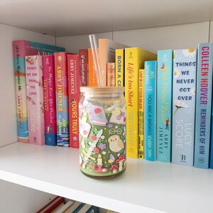 May include: A glass jar with a wooden lid and a glass straw, decorated with whimsical illustrations of books, flowers, and an owl. The jar is on a white shelf with a collection of colorful books.