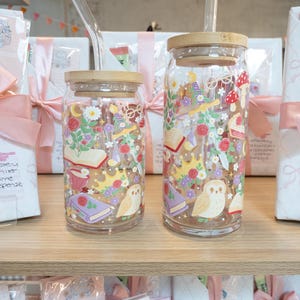 May include: Two glass drinking jars with bamboo lids and clear glass straws. The jars are decorated with colorful illustrations of books, flowers, crowns, and owls. The jars are on a wooden shelf with wrapped gifts in the background.