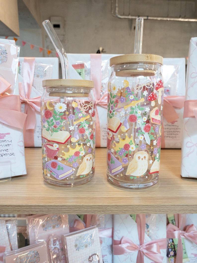 May include: Two glass drinking jars with bamboo lids and clear glass straws. The jars are decorated with colorful illustrations of books, flowers, crowns, and owls. The jars are on a wooden shelf with wrapped gifts in the background.