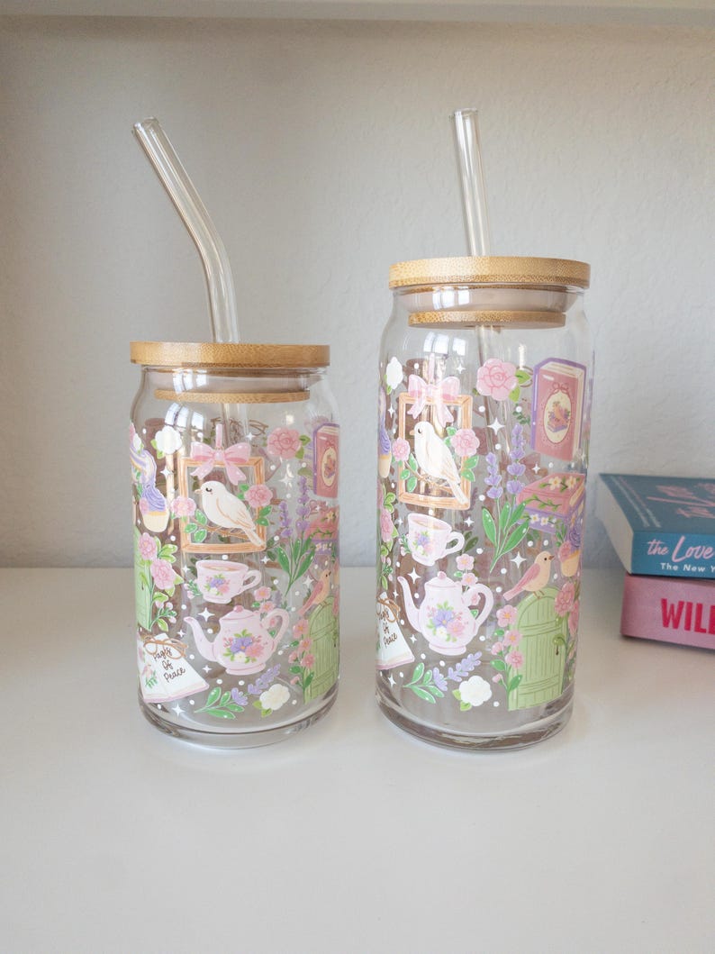 May include: Two clear glass drinking cups with bamboo lids and glass straws. The cups feature a floral design with teacups, teapots, birds, and the text "People of Peace". The design includes pink, purple, and green elements.