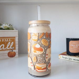 May include: A clear glass tumbler with a bamboo lid and a clear straw, filled with a light brown beverage. The tumbler is decorated with fall-themed illustrations, including pie, sweaters, and leaves. A wooden box with the text "FALL" is visible.