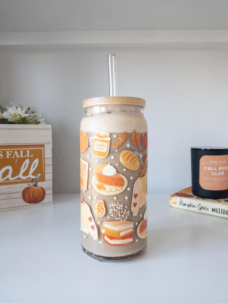 May include: A clear glass tumbler with a bamboo lid and a clear straw, filled with a light brown beverage. The tumbler is decorated with fall-themed illustrations, including pie, sweaters, and leaves. A wooden box with the text "FALL" is visible.