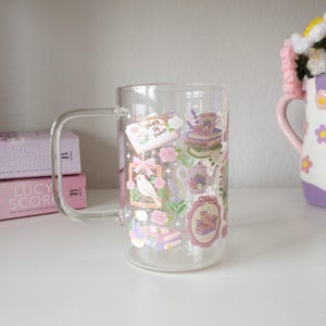 May include: A clear glass mug with a handle, decorated with floral and book-themed stickers in pink, purple, and green. The stickers include a bird, teapot, and the text "Pages of Peace".