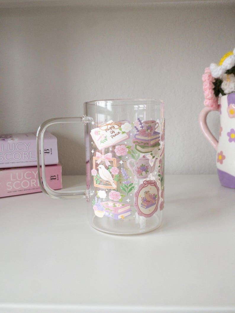May include: A clear glass mug with a handle, decorated with floral and book-themed stickers in pink, purple, and green. The stickers include a bird, teapot, and the text "Pages of Peace".