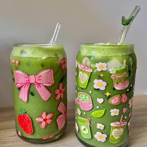 以下が含まれることがあります： 緑色の抹茶が入った2つの透明なガラス製タンブラーで、それぞれにガラス製のストローが付いています。左側のグラスには大きなピンクのリボンとイチゴのデカールがあり、右側のグラスには抹茶カップ、ピンクの花、ハートの小さなステッカーが貼られています。