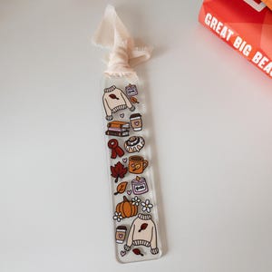 May include: A clear acrylic bookmark with a cream-colored ribbon. The bookmark features fall-themed illustrations, including a sweater, books, coffee cups, a cinnamon roll, and a pumpkin. The word "Fall" is also visible.