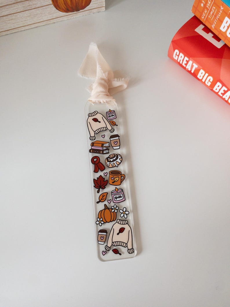 May include: A clear acrylic bookmark with a cream-colored ribbon. The bookmark features fall-themed illustrations, including a sweater, books, coffee cups, a cinnamon roll, and a pumpkin. The word "Fall" is also visible.