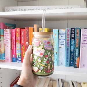 May include: A glass tumbler with a bamboo lid and a clear straw, filled with a green beverage. The tumbler is decorated with a floral and book-themed design. The background features a bookshelf filled with colorful books.