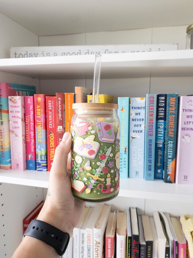 May include: A glass tumbler with a bamboo lid and a clear straw, filled with a green beverage. The tumbler is decorated with a floral and book-themed design. The background features a bookshelf filled with colorful books.