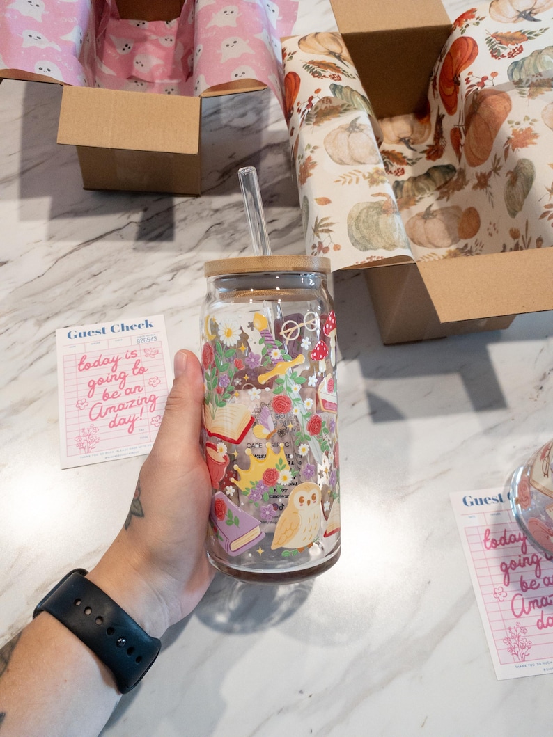 May include: A clear glass tumbler with a bamboo lid and a straw. The tumbler is decorated with a whimsical floral and owl design. The words "today is going to be an Amazing day!" are on a pink guest check.