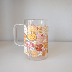May include: A clear glass mug with a handle, decorated with a repeating pattern of baked goods in shades of yellow, pink, and brown. The design includes croissants, hearts, and other pastries, with small white flower accents.
