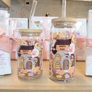May include: Two clear glass tumblers with bamboo lids and glass straws. The tumblers feature a Halloween-themed design with a haunted house, pumpkins, and the text "HAUNTED BOOKS". The design includes a ghost reading a book.