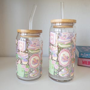 May include: Two clear glass tumblers with bamboo lids and glass straws. The tumblers are decorated with pastel illustrations of books, teacups, birds, and flowers. The tumblers are of different sizes.