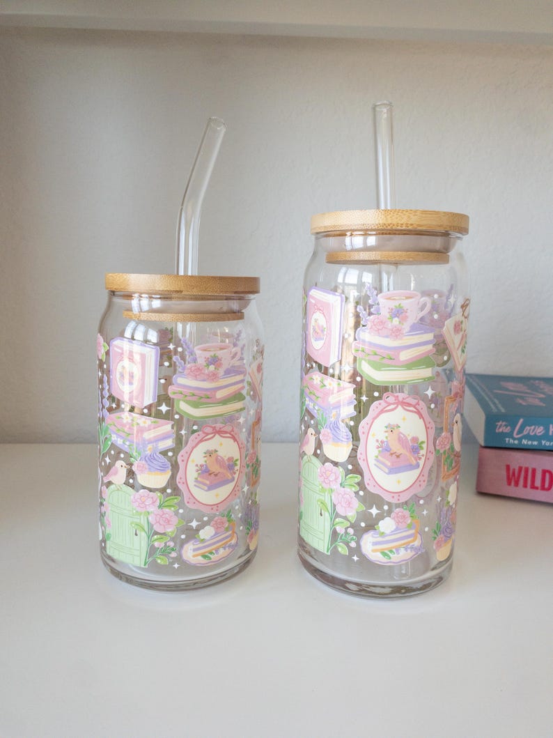 May include: Two clear glass tumblers with bamboo lids and glass straws. The tumblers are decorated with pastel illustrations of books, teacups, birds, and flowers. The tumblers are of different sizes.