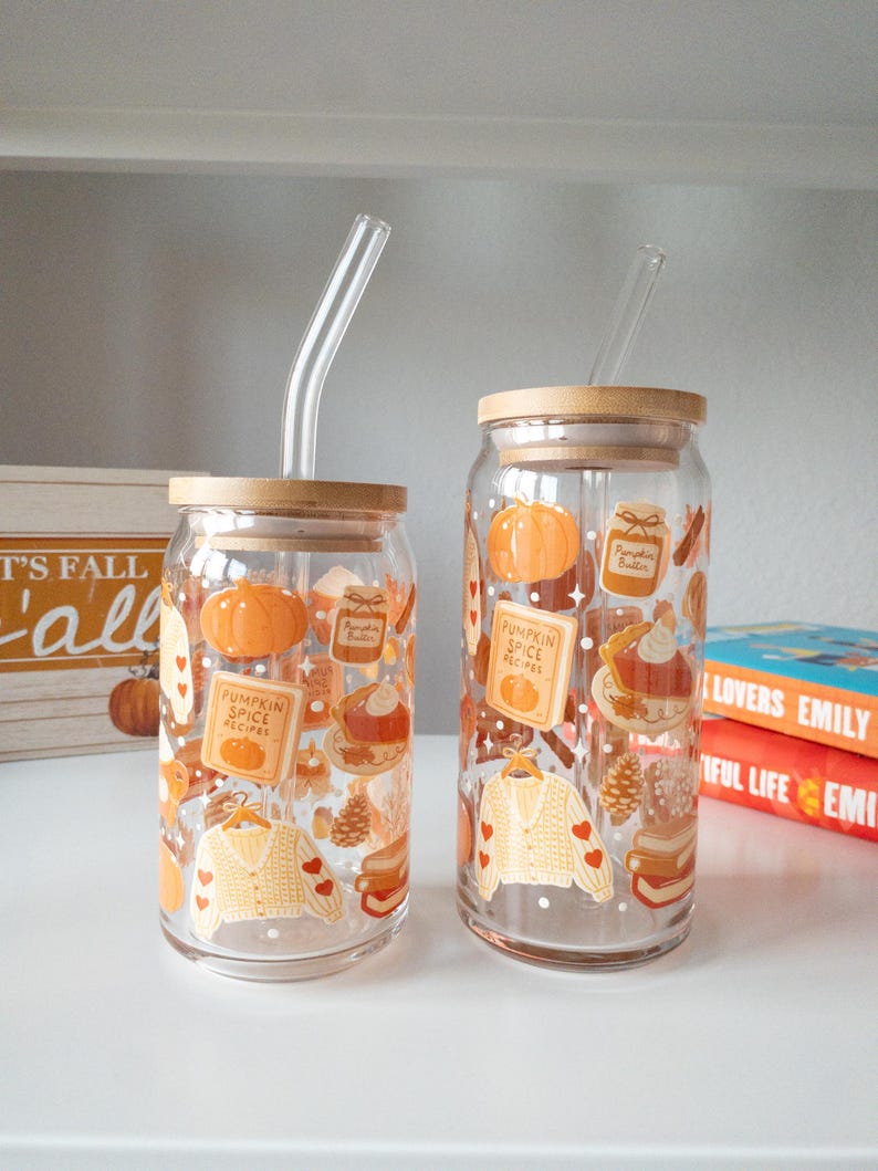 May include: Two clear glass tumblers with bamboo lids and glass straws. The tumblers are decorated with fall-themed illustrations, including pumpkins, sweaters, and the text "Pumpkin Spice Recipes".