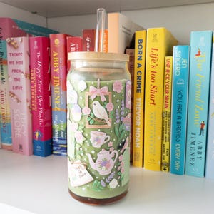 May include: A glass tumbler with a bamboo lid and a clear straw, decorated with a floral and bird design in shades of green, pink, and white. The tumbler is filled with a green beverage and sits on a white shelf with colorful books.