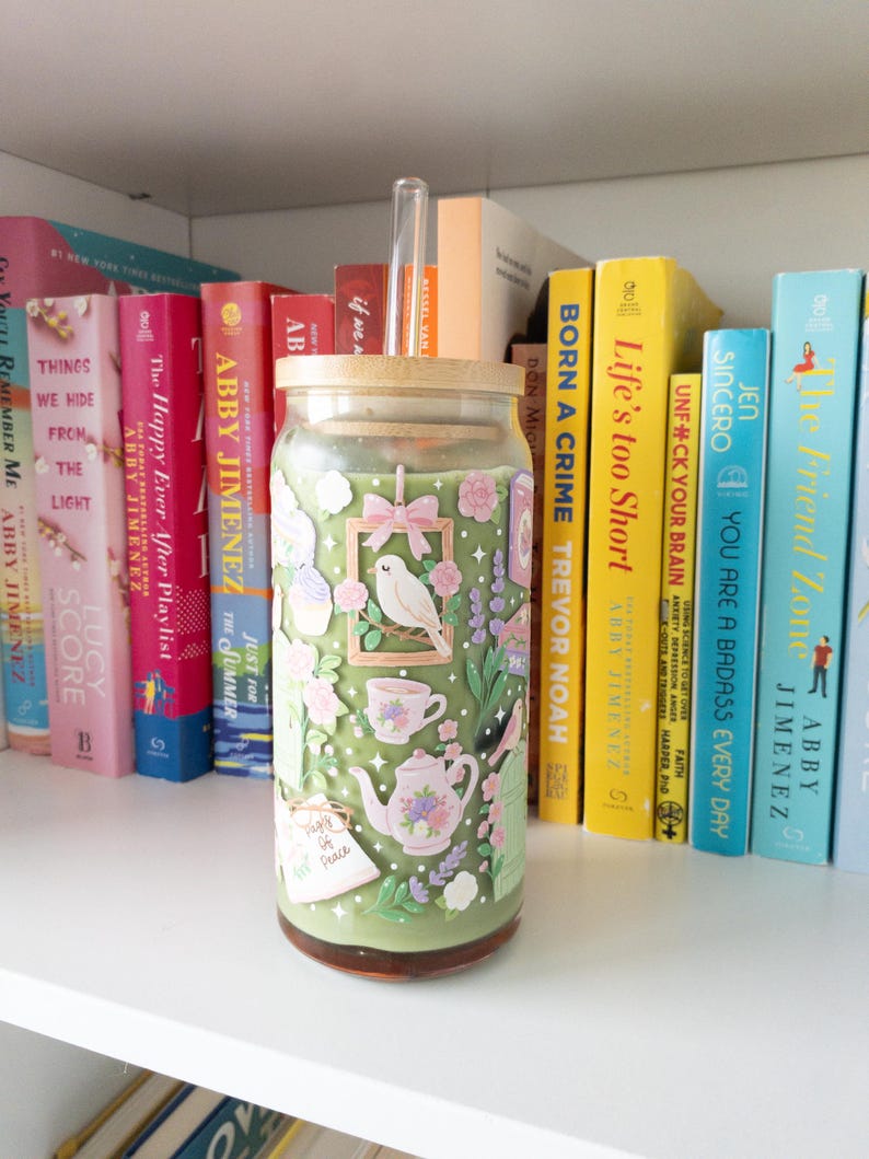 May include: A glass tumbler with a bamboo lid and a clear straw, decorated with a floral and bird design in shades of green, pink, and white. The tumbler is filled with a green beverage and sits on a white shelf with colorful books.