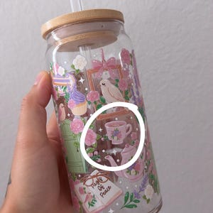 May include: A clear glass tumbler with a bamboo lid and a clear straw. The tumbler is decorated with a floral and bird design, including a teacup and teapot. The words "Pages of Peace" are visible on a book.