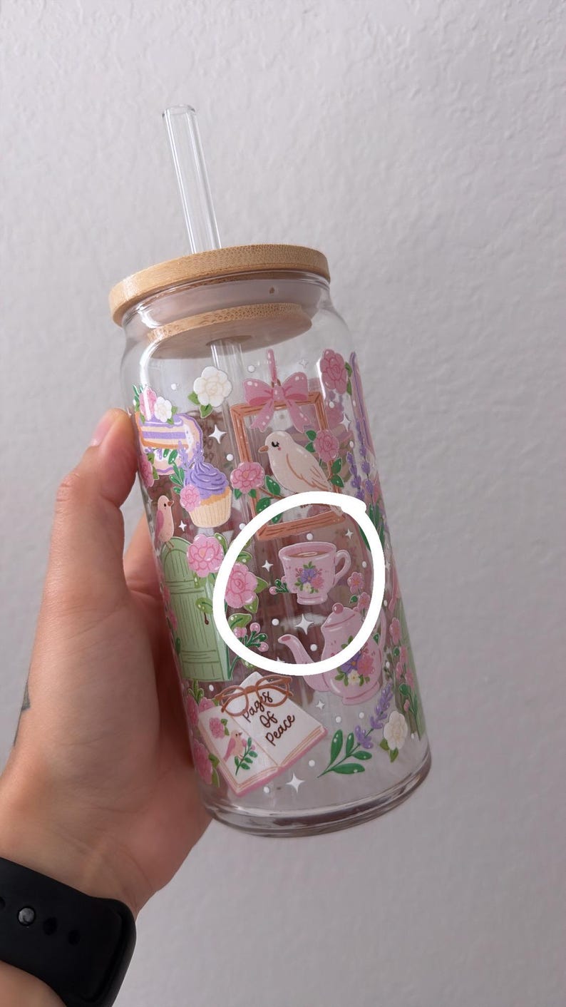 May include: A clear glass tumbler with a bamboo lid and a clear straw. The tumbler is decorated with a floral and bird design, including a teacup and teapot. The words "Pages of Peace" are visible on a book.