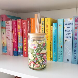 May include: A decorative glass jar with a wooden lid, featuring a whimsical design of flowers, mushrooms, and other illustrations. The jar is displayed on a white shelf with a collection of colorful books.