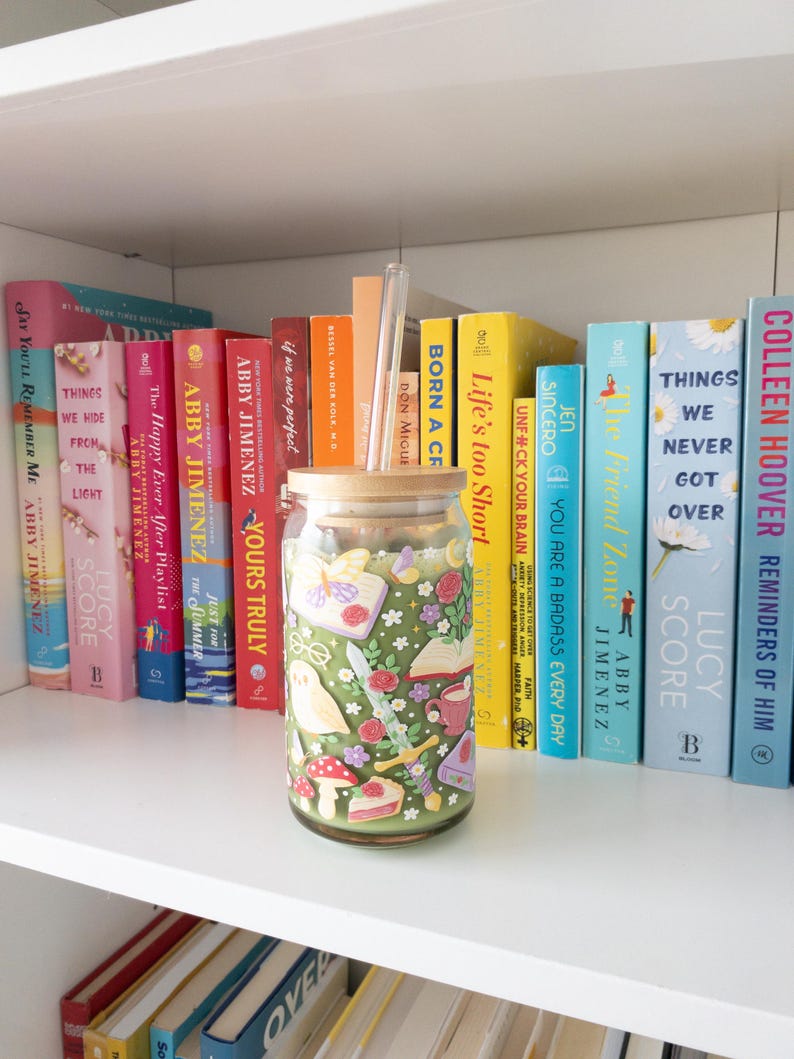 May include: A decorative glass jar with a wooden lid, featuring a whimsical design of flowers, mushrooms, and other illustrations. The jar is displayed on a white shelf with a collection of colorful books.