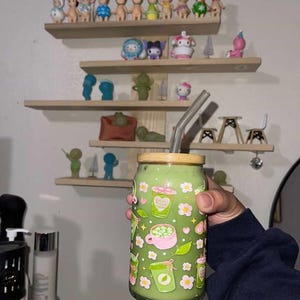 以下が含まれることがあります： 竹製の蓋とガラス製のストローが付いた、緑色の抹茶飲料が入った透明なガラス製タンブラー。グラスの外側には、ピンクと緑のドリンクカップ、小さな白い花、ピンクのハートが繰り返されるパターンで装飾されています。