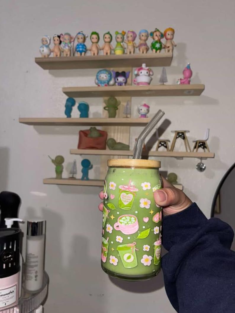 以下が含まれることがあります： 竹製の蓋とガラス製のストローが付いた、緑色の抹茶飲料が入った透明なガラス製タンブラー。グラスの外側には、ピンクと緑のドリンクカップ、小さな白い花、ピンクのハートが繰り返されるパターンで装飾されています。