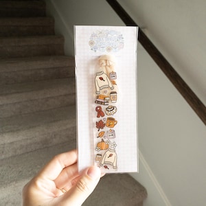 May include: A clear bookmark with fall-themed illustrations, including a sweater, pumpkin, coffee cup, and leaves. The bookmark is held in a hand and is packaged in a clear plastic sleeve. The text "Stay Bold" is visible at the top.