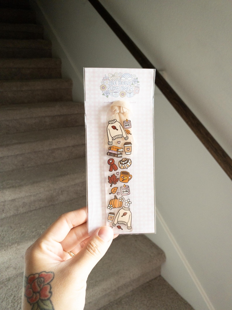 May include: A clear bookmark with fall-themed illustrations, including a sweater, pumpkin, coffee cup, and leaves. The bookmark is held in a hand and is packaged in a clear plastic sleeve. The text "Stay Bold" is visible at the top.