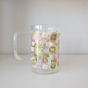 以下が含まれることがあります： 緑茶のカップ、ピンクの花、小さなハート、スライスされたイチゴなど、抹茶をテーマにしたイラストが繰り返し描かれた、取っ手付きの透明なガラス製マグカップ。緑、ピンク、白、黄色の色合いでデザインされています。