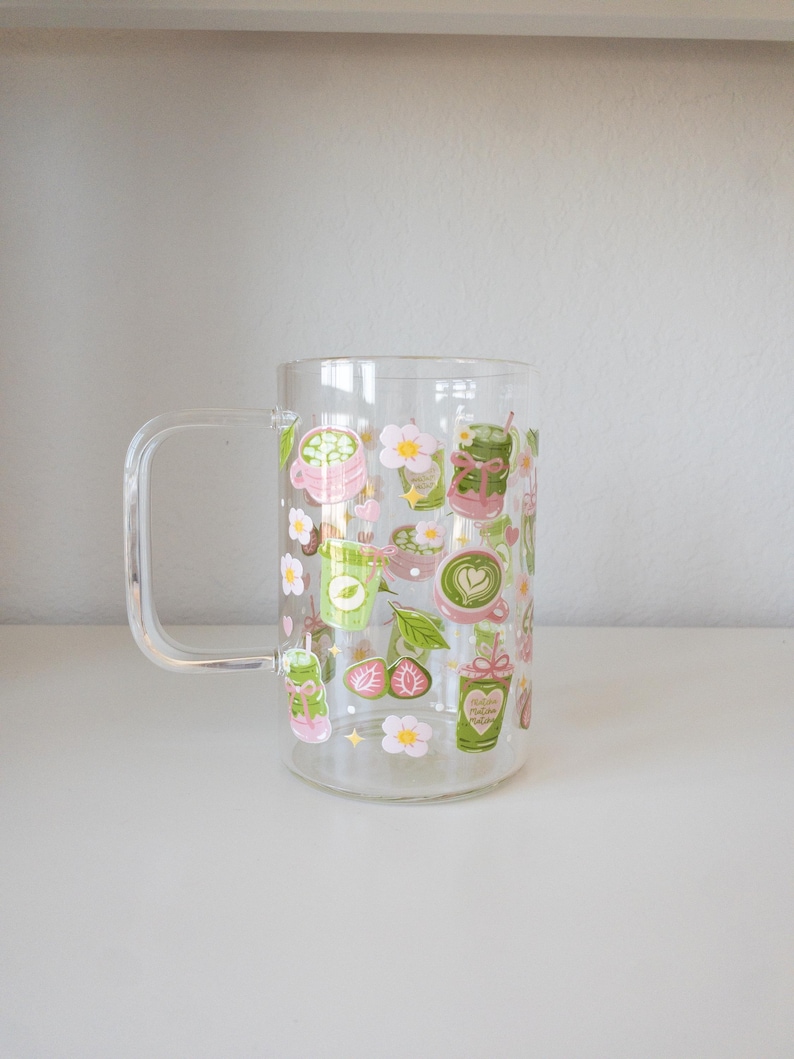 以下が含まれることがあります： 緑茶のカップ、ピンクの花、小さなハート、スライスされたイチゴなど、抹茶をテーマにしたイラストが繰り返し描かれた、取っ手付きの透明なガラス製マグカップ。緑、ピンク、白、黄色の色合いでデザインされています。