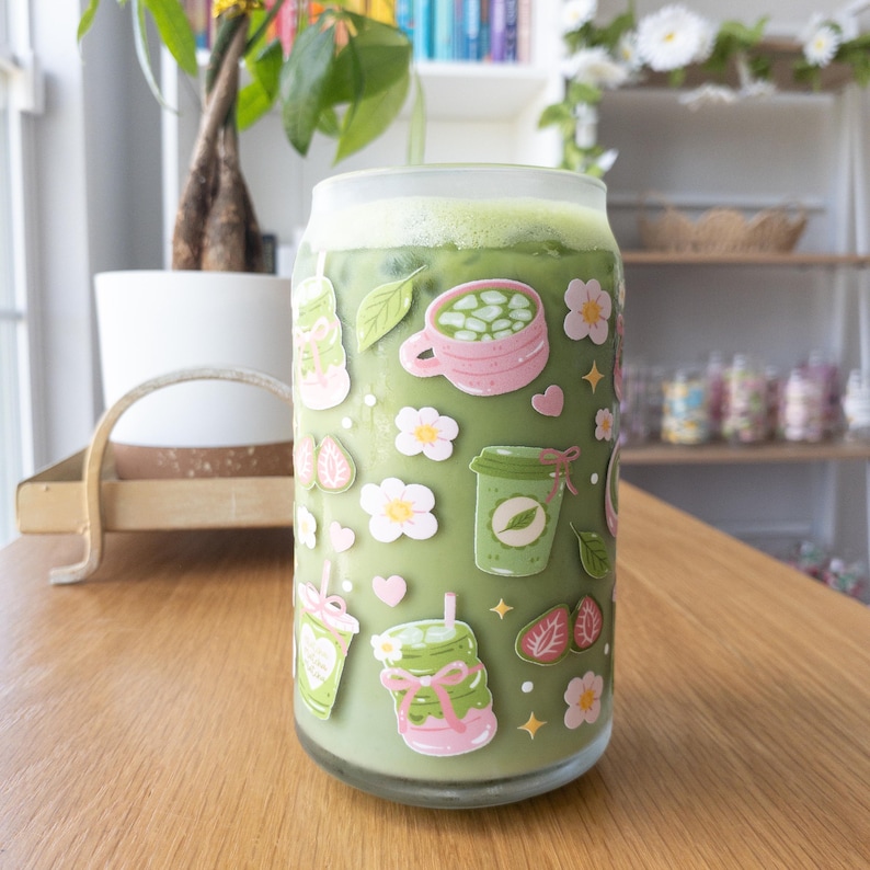 以下が含まれることがあります： 薄緑色の抹茶ラテが入った、缶の形をしたすりガラス製のタンブラー。外側には、ピンクの花、緑のティーカップ、スライスされたイチゴ、小さなハート、黄色い星などが描かれたカラフルなステッカーが貼られています。