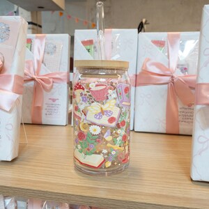 May include: A clear glass tumbler with a bamboo lid and a glass straw. The tumbler is decorated with a colorful floral and butterfly design. The tumbler is sitting on a wooden shelf, with gift boxes in the background.