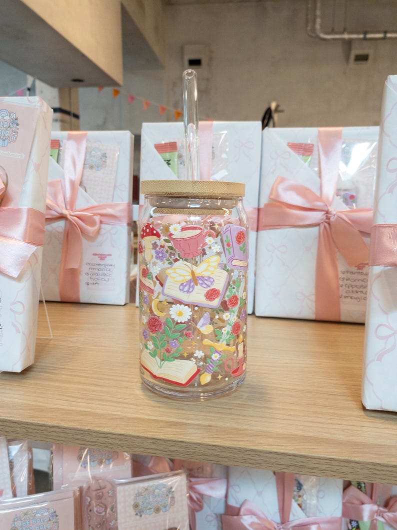 May include: A clear glass tumbler with a bamboo lid and a glass straw. The tumbler is decorated with a colorful floral and butterfly design. The tumbler is sitting on a wooden shelf, with gift boxes in the background.
