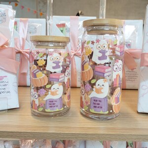 May include: Two clear glass tumblers with bamboo lids and glass straws. The tumblers feature a whimsical design of a ghost reading a book, pumpkins, and fall leaves. The text "Read Till The End... And After" is printed on the bottom.